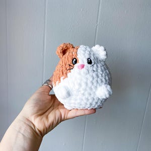 May include: A crocheted plush toy shaped like a guinea pig. The toy is white with a brown patch on one side. It has black eyes, a pink nose, and a stitched-on smile.
