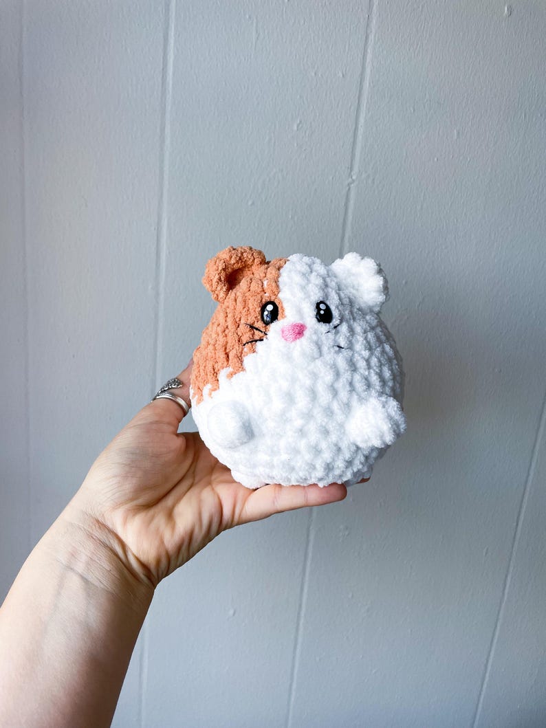 May include: A crocheted plush toy shaped like a guinea pig. The toy is white with a brown patch on one side. It has black eyes, a pink nose, and a stitched-on smile.