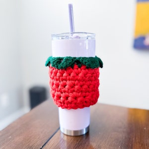 May include: A white tumbler with a red and green crocheted cup cozy. The cozy is shaped like a strawberry. A purple straw is sticking out of the tumbler.