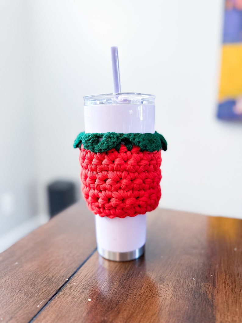 May include: A white tumbler with a red and green crocheted cup cozy. The cozy is shaped like a strawberry. A purple straw is sticking out of the tumbler.