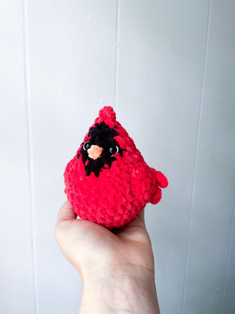May include: A crocheted cardinal bird plush toy with black eyes and a bright red body. The toy is held in a person's hand.