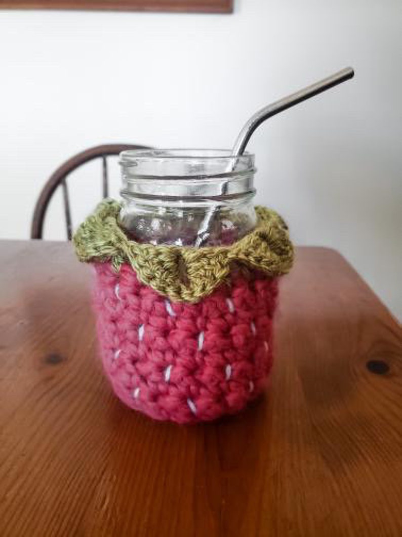 May include: A crocheted strawberry-shaped glass cozy with green leaves, a clear glass jar, and a metal drinking straw.