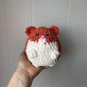 May include: A crocheted plush toy shaped like a hamster. The hamster is orange and white with black embroidered eyes and a pink nose.