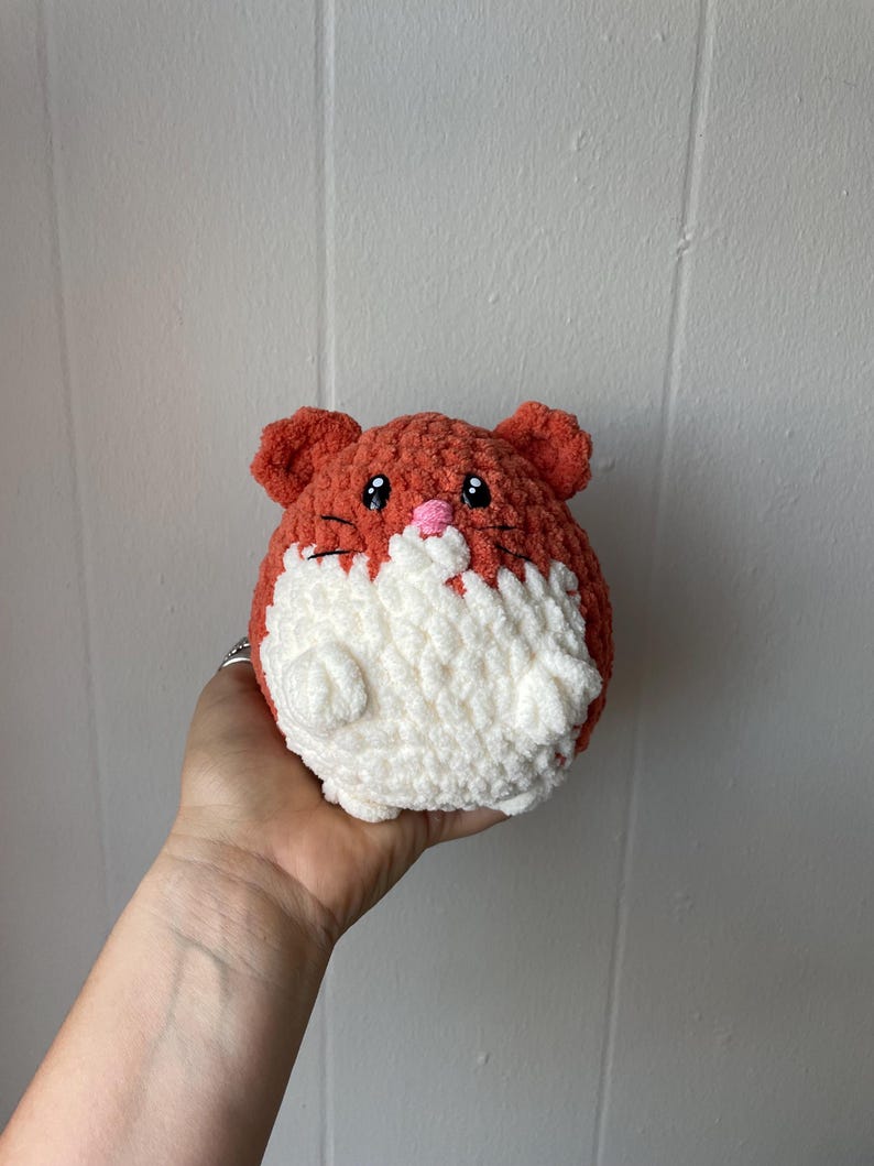 May include: A crocheted plush toy shaped like a hamster. The hamster is orange and white with black embroidered eyes and a pink nose.