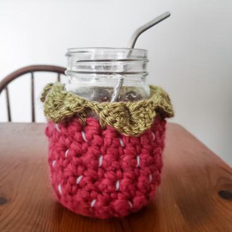May include: A crocheted strawberry-shaped cozy for a glass jar, with a green crocheted top and a red body. The cozy is on a wooden table with a metal straw sticking out of the jar.