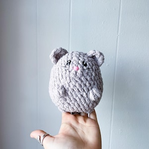 May include: A crocheted plush toy shaped like a mouse. The mouse is light gray with pink accents for the nose and eyes.