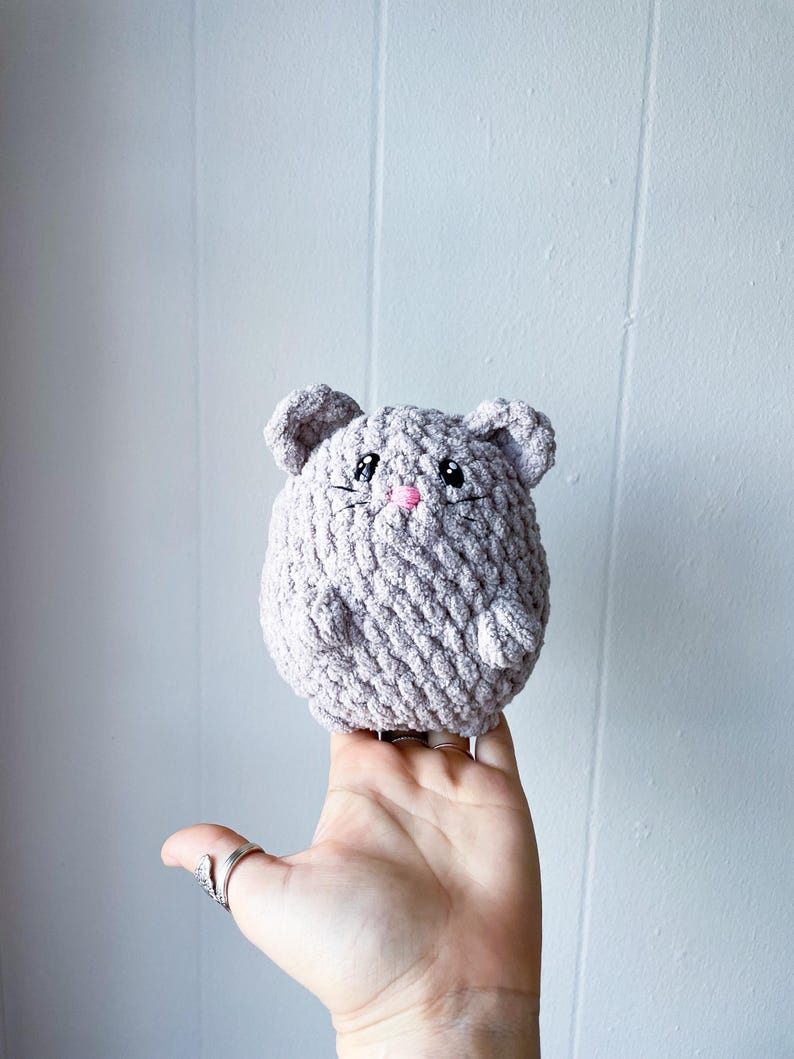 May include: A crocheted plush toy shaped like a mouse. The mouse is light gray with pink accents for the nose and eyes.