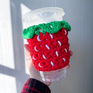 May include: A red and white crocheted strawberry-shaped cup cozy with green leaves. The cozy is on a clear plastic cup.