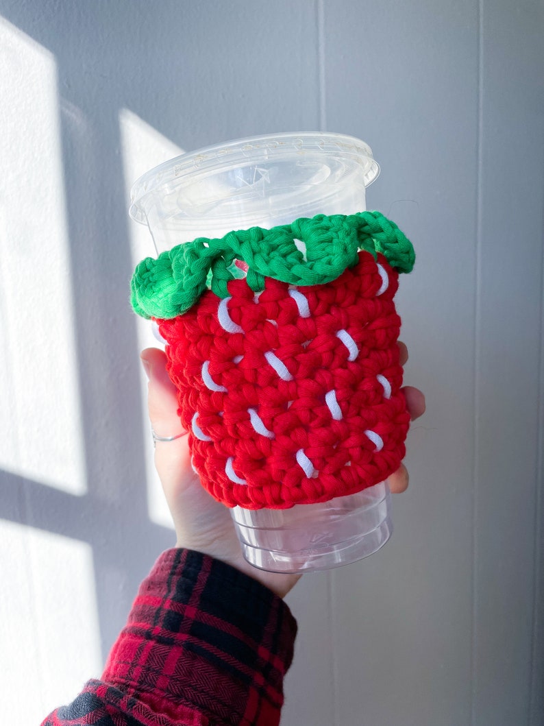 May include: A red and white crocheted strawberry-shaped cup cozy with green leaves. The cozy is on a clear plastic cup.