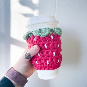 May include: A red and white crocheted coffee cup cozy shaped like a strawberry with green leaves.