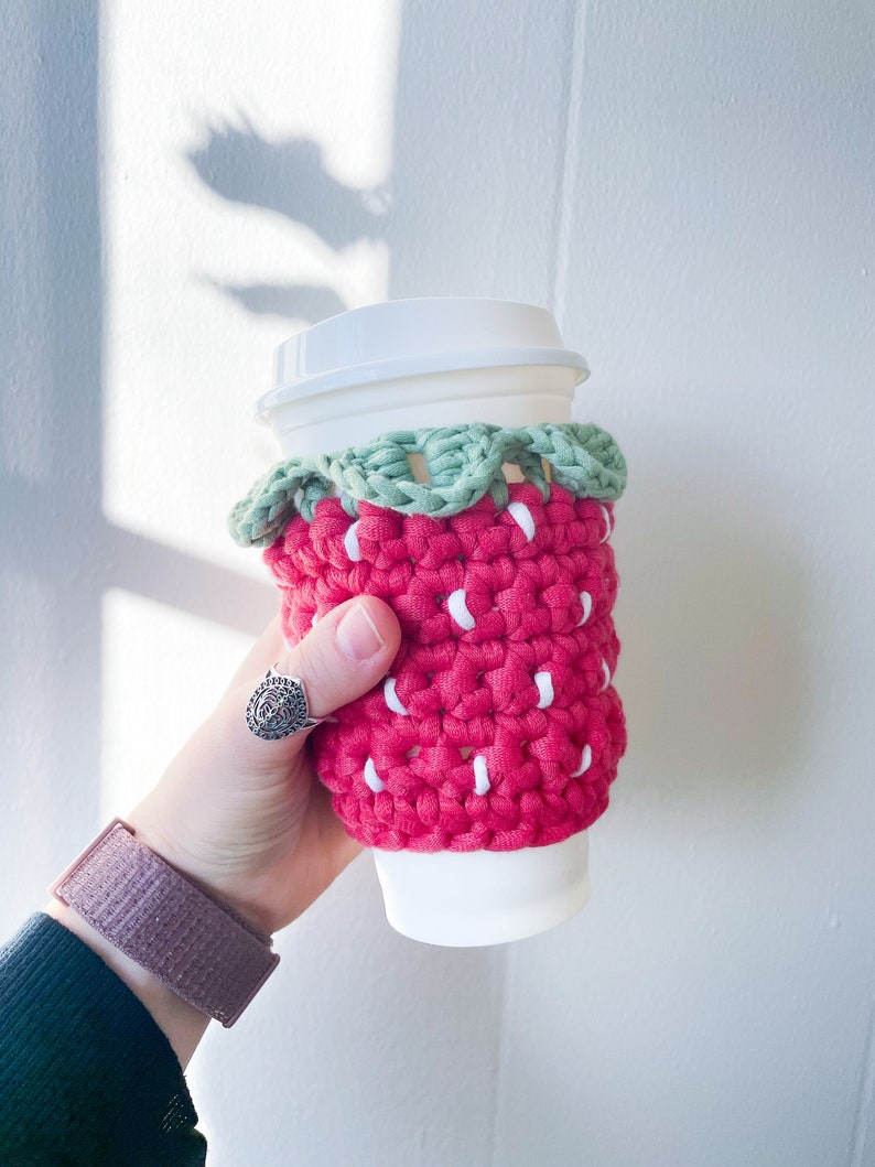 May include: A red and white crocheted coffee cup cozy shaped like a strawberry with green leaves.