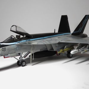 Könnte beinhalten: Ein graues und schwarzes Flugzeugmodell mit einem blauen und weißen Streifen an der Seite. Das Flugzeug hat sein Fahrwerk ausgefahren und trägt mehrere Raketen. Das Flugzeug trägt die Nummer "00" an der Seite.