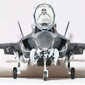 May include: A detailed model of a dark gray fighter jet with open cockpit. The jet has white accents on the nose and landing gear. The wings are extended, and the jet is positioned on a white surface.