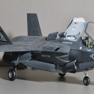 May include: A detailed model of a dark gray F-35 fighter jet with open cockpit and landing gear down. The model has intricate details, including markings and a star emblem. The wings and tail fins are visible, and the model is set against a neutral background.