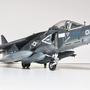 May include: A dark gray model airplane with the number "02" on the side. The model has a clear cockpit, gray undercarriage, and gray external fuel tanks. The wings have missiles attached. The model is on a white surface.