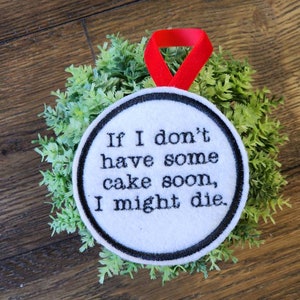 May include: A white circular ornament with black text that reads "If I don't have some cake soon, I might die." The ornament has a black border and a red ribbon for hanging. It is set against a backdrop of green foliage.