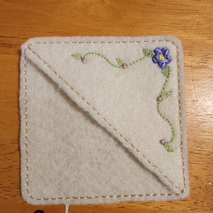 Può includere: Un sottobicchiere quadrato in feltro bianco sporco con un ricamo floreale. Il ricamo presenta un fiore blu con centro giallo, tralci verdi e accenti rosa. Il sottobicchiere ha un bordo cucito e una cucitura diagonale.