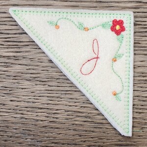 May include: A cream-colored, triangular felt applique with a decorative floral design and the letter "J" embroidered in red. The edges are stitched in green, and the floral design includes a red flower and orange accents.