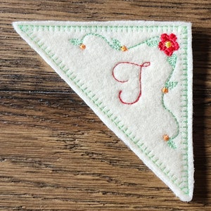 May include: A triangular, off-white felt applique with a green stitched border. It features a red embroidered flower, green vines, orange accents, and a red letter "J". The applique is on a wooden surface.