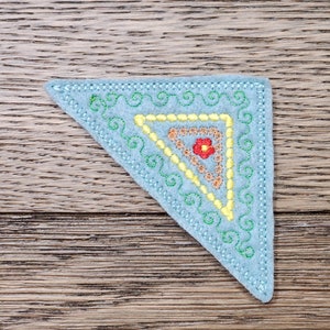 May include: A light blue felt triangle with embroidered details. The design features a yellow and orange inner triangle, a red flower, and green swirls. The edges are stitched with a light blue thread. The item is placed on a wooden surface.