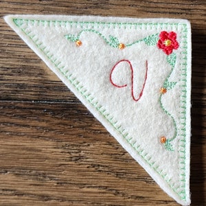 Könnte beinhalten: Ein weißes Eck-Lesezeichen aus Filz mit grün genähten Kanten und dem rot gestickten Buchstaben "V". Das Lesezeichen hat ein Blumendesign mit grünen Blättern, orangefarbenen Punkten und einer roten Blume.