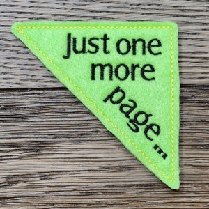 Puede incluir: Un marcapáginas triangular de fieltro verde lima con el texto "Just one more page..." bordado en negro. Los bordes están delineados con costuras amarillas. El marcapáginas está sobre una superficie de madera.