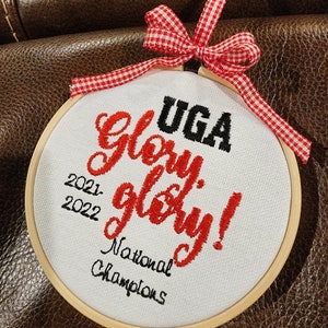 May include: A white fabric hoop with the text "UGA Glory Glory! National Champions 2021-2022" embroidered in red and black thread. A red and white gingham ribbon is tied at the top.