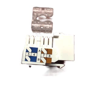 Puede incluir: Un conector RJ45 blanco con diagramas de cableado codificados por colores azul y marrón, y un clip de plástico transparente. El conector tiene pines numerados para conexiones de red.