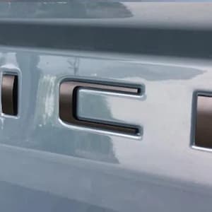 May include: Close-up of a vehicle's tailgate with the word "RICK" in black, three-dimensional lettering. The letters are slightly raised and have a metallic sheen, contrasting against the light blue surface.