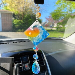 May include: A car rearview mirror charm featuring a blue, green, and yellow glass mosaic with a sun and wave design. The charm is attached to a silver chain with crystal beads.