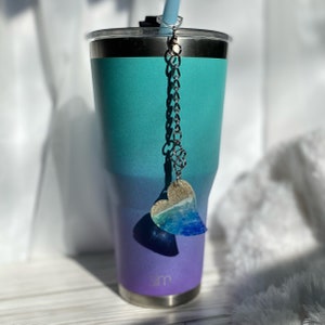 May include: A teal and purple stainless steel tumbler with a silver chain and a heart-shaped charm. The charm is made of clear resin with blue and white swirls, resembling ocean waves.