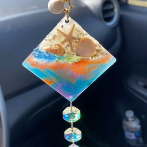 May include: A diamond-shaped resin charm with a beach scene, including a starfish, seashells, and sand. The charm is attached to a gold chain and hangs from three crystal beads.