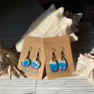 May include: A pair of ocean wave themed earrings with a blue, purple, and white color scheme. The earrings are made of resin and have a silver hook. The earrings are displayed on a brown card with the text "Blessed" and "Greater Expectancy For You".