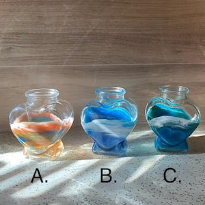 May include: Three clear glass heart-shaped vases filled with colorful sand. The sand in each vase is a different color combination: orange and yellow, blue and white, and teal and white. Each vase is labeled with a letter: A, B, and C.