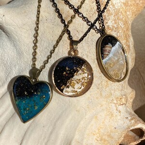 May include: Three gold necklaces with different pendants. The first pendant is a heart shape with a black and blue resin filled with gold flakes. The second pendant is a round circle with a black and white resin filled with gold flakes. The third pendant is an oval with a clear resin filled with gold flakes and a small seashell.