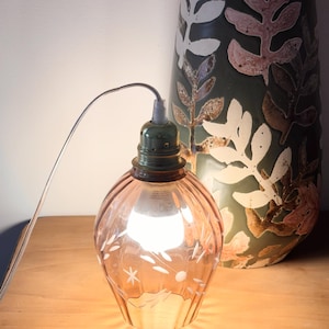 Peut inclure: Lampe de table vintage en verre rose, ornée de motifs floraux et étoilés gravés. La lampe a une base verte et une ampoule visible. Un grand vase vert avec des motifs de feuilles blanches et roses est en arrière-plan.