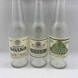 Puede incluir: Tres botellas de vidrio transparente con tapones de corcho y etiquetas. Las botellas están llenas de un líquido transparente. Las etiquetas están en diferentes idiomas y presentan diseños diferentes. La primera botella está etiquetada "ПШЕНИЧНАЯ ВОДКА" (vodka de trigo) y tiene una etiqueta roja y blanca. La segunda botella está etiquetada "VIIN" y tiene una etiqueta verde y blanca. La tercera botella está etiquetada "VIIN VODKA" y tiene una etiqueta verde y blanca.