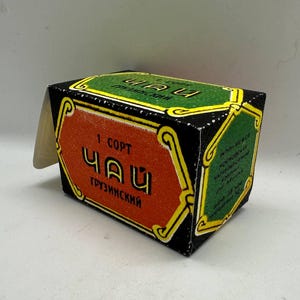 Könnte beinhalten: Eine Vintage-Teebox mit schwarzem Hintergrund und grünen und orangefarbenen Paneelen. Die Box zeigt kyrillischen Text und goldfarbene dekorative Elemente. Eine weiße Papierlasche ragt von oben heraus.