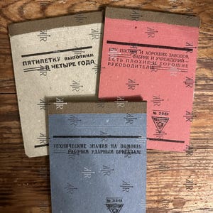Op de afbeelding: Drie vintage notitieboekjes met Russische tekst. De notitieboekjes zijn beige, rood en blauw. Elk notitieboekje heeft een andere kleur en tekst. De notitieboekjes zijn rechthoekig met een bruine binding.