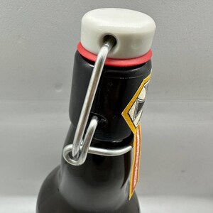 May include: A dark brown glass bottle with a white ceramic stopper, red rubber seal, and a metal swing top closure. The bottle has a small label with yellow and red accents. The bottle is designed for beverages.