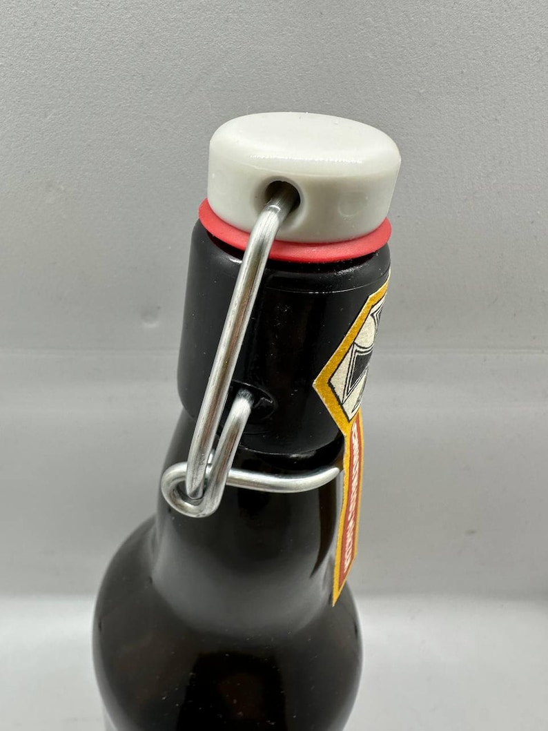 May include: A dark brown glass bottle with a white ceramic stopper, red rubber seal, and a metal swing top closure. The bottle has a small label with yellow and red accents. The bottle is designed for beverages.