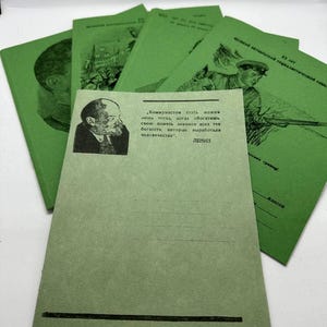 May include: A collection of green booklets with black text and illustrations. One booklet features a portrait of a man and text in Russian. Other booklets show illustrations of soldiers and portraits. The booklets are stacked on a white surface.