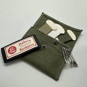 May include: An olive green sewing kit with spools of thread in white and green shades, safety pins, and a label with red text that reads "Nadeln für unsere Soldaten". The kit is made of canvas.