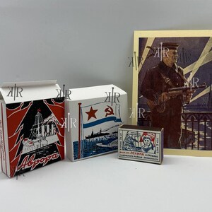 May include: A collection of vintage matchboxes and a postcard with Soviet-era designs. The matchboxes depict ships, flags, and a soldier. The postcard shows a soldier with a weapon. The colour scheme includes red, white, and blue.