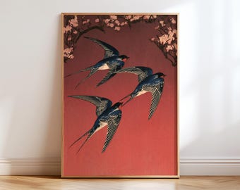 May include: A framed wall art print featuring three swallows in flight against a solid deep red background. The birds have dark blue and black plumage with white underbellies. Pale pink cherry blossom branches frame the top corners.