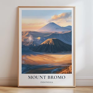 May include: A vertical wall art print in a light wood frame featuring a landscape illustration of Mount Bromo in Indonesia. The scene depicts volcanic peaks under a soft sunrise sky with golden light hitting the slopes and dark blue shadows below.
