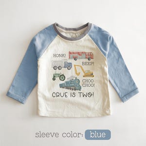 May include: A long-sleeved, raglan-style shirt with blue sleeves and a cream-colored body. The shirt features illustrations of vehicles like a truck, tractor, and train, with the text "CRUE IS TWO!" and vehicle sound effects.