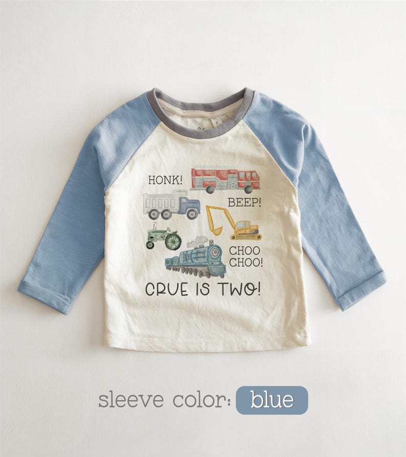 May include: A long-sleeved, raglan-style shirt with blue sleeves and a cream-colored body. The shirt features illustrations of vehicles like a truck, tractor, and train, with the text "CRUE IS TWO!" and vehicle sound effects.