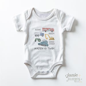 May include: White baby onesie with colorful illustrations of a truck, fire truck, tractor, excavator, and train. Text includes "HONK!", "BEEP!", "CHOO CHOO!", and "KADEN IS TWO!". The onesie has a snap closure.