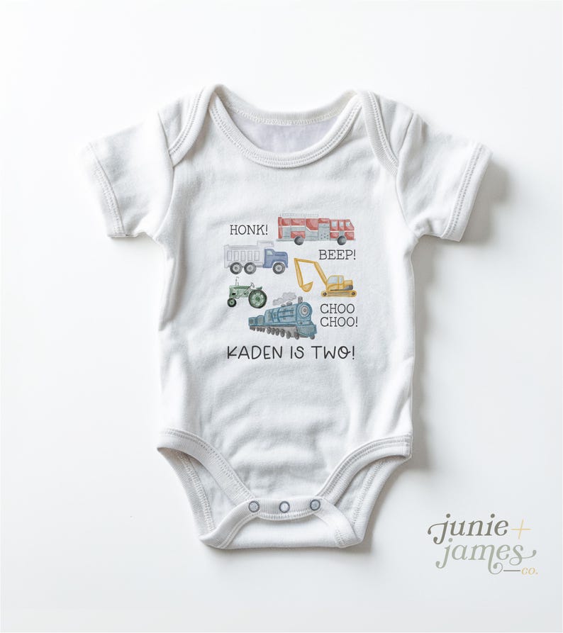 May include: White baby onesie with colorful illustrations of a truck, fire truck, tractor, excavator, and train. Text includes "HONK!", "BEEP!", "CHOO CHOO!", and "KADEN IS TWO!". The onesie has a snap closure.